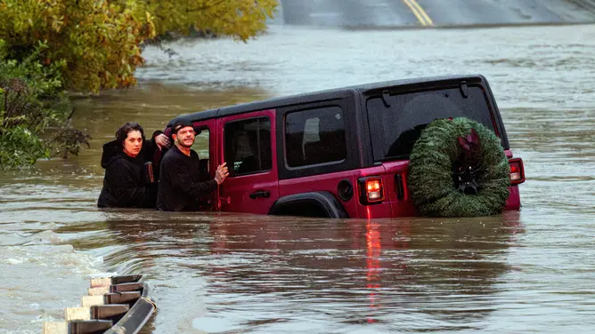 A stranded couple hold onto their vehicle that became stuck in flood water in Windsor, California on November 22, 2024. A "bomb cyclone" was lashing the western United States bringing hurricane-force winds that have killed at least two people and left half a million without power. (Photo by JOSH EDELSON / AFP) (Photo by J via Getty Images) (JOSH EDELSON/AFP)