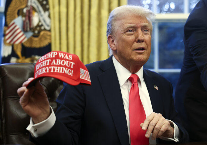 President Donald Trump holds up a hat as he talks with reporters in the Oval Office at the White House in Washington, Tuesday, Feb. 25, 2025. (Pool via AP)