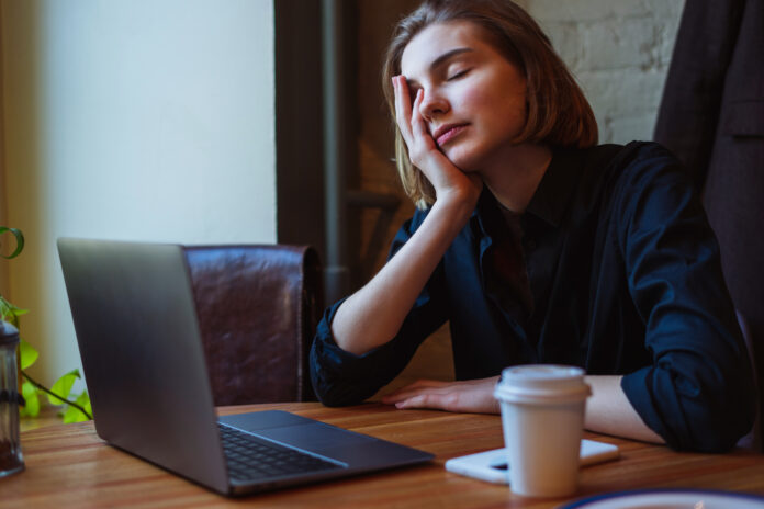 A tired young woman in a shirt sitting in a cafe, rubbing her eyes after a long day working on her laptop. A disposable coffee cup and phone lay on the table. Daylight from the window.