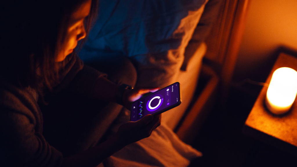 Young woman using healthcare app on smartphone to improve sleep while sitting on bed, getting ready to sleep. Healthcare and technology.
